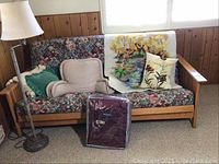 Wooden futon sofa bed with floral cushions, satin bedspread in packaging, needlework butterfly pillow, reading backrest pillow, and floor lamp beside the sofa
