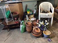 Photo showing a variety of garden supplies including terracotta pots, soil bag, garden hose on a wire rack, extension cord, three white plastic stacking chairs, a rusty metal wheelbarrow, plant stakes, and a garden work stool.
