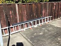 Full view of aluminum extension ladder laid against a wooden fence with the black flexible drain pipe on top, showing overall condition and length.