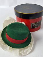 Miniature green felt hat with red fabric band displayed in front of a round red and black box labeled 'Resista', with white tissue paper.