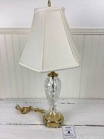 Full view of the lamp showing crystal body, brass base, and white fabric shade.