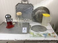 Photo of assorted vintage kitchen aluminum pieces, glass pitcher, small jar with wooden lid, colorful rectangular glass dish, and red Ice-O-Mat hand-crank crushed ice maker.
