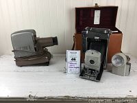 Photo showing vintage TDC projector in brown, Polaroid 150 camera, Wink flash light, and brown leather carrying case on white surface.