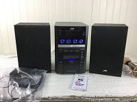 Front view of JVC micro component system with main unit between two speakers, including power cord and instructions card.