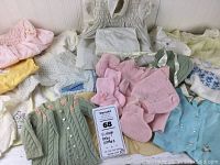 Various vintage baby clothes laid out including knitted sweaters, booties, a christening gown and cap, assorted tops in pastel colors.