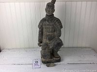 Full view of dark clay warrior statue kneeling on one knee, detailed armor and topknot hairstyle visible. Statue approximately 20.5 inches tall, standing on rectangular base. One hand missing, damage visible.