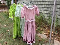 Photo showing three dresses hanging outdoors on a rack including green, white floral, and pink floral dresses.