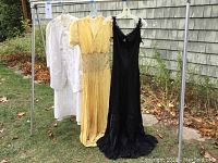 Full view of three vintage dresses hanging on rack outside showing white, yellow, and black dresses.
