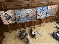 Photo shows three carved wooden duck decoys on the carpet placed in front of four wooden TV trays with painted duck scenes. Trays are supported by folding legs and displayed upright against wood paneled wall.