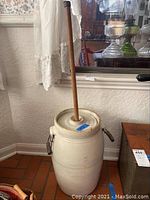 Full view of tall antique stoneware butter churn with wooden dasher inserted in lid. Metal side handles visible.