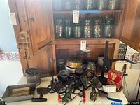 Wide view of vintage kitchen items on table in front of wooden cabinet showcasing jars, utensils, molds, and bottle capper.
