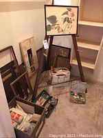 Overall view of easel with framed print, several picture frames, paint and supplies boxes on floor.