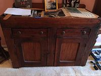 Front view of the antique dry sink cabinet showing two drawers with round wooden knobs and two paneled doors below, with several newspapers and items on top.