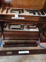Wide view showing various vintage drawing and carpentry tools arranged on and near a wooden desk, including framing squares, hand saws, levels, and mallet.