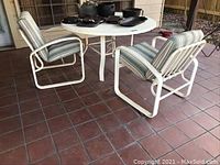 Patio table with four chairs arranged around it on tiled porch floor, white metal frames, cushions have green and white stripes. Weathered condition visible on frames and cushions.