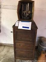 Wood cabinet with dark finish, standing on floor with box of Victrola parts on top. Visible wear and broken hinges on lid.
