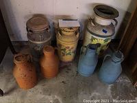 Seven milk cans of different sizes and paint styles arranged on floor against a wall including painted floral designs and plain cans