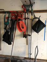 Wide view showing the hanging yard tools: rake with green handle, orange extension cord on reel, Black & Decker blower, Gilmore green sprinkler, pole saw, garden hoe