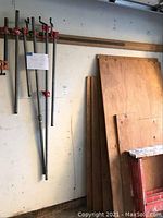 Pipe clamps hanging on wall next to plywood sheets and saw horses in garage or workshop setting.