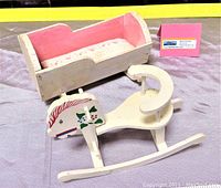 Photo of wooden rocking horse and bassinet doll furniture. Rocking horse decorated with floral designs on the horse's head and painted cream with a seating safety bar. Bassinet painted cream with pink interior and floral bedding fabric.