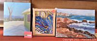 Three paintings together showing the snowy mountain cabin, abstract colorful painting, and rocky coastal scene.