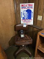Photo of vintage brown rolling desk chair with Rolleicord box camera placed on seat, and Ike & Tina Turner poster pinned to wood panel wall behind.