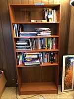 Full view of tall wooden bookcase filled with books on multiple shelves, organized with labeled categories, against wood paneled wall.