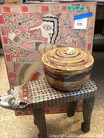 Decorative wooden cat-shaped stool painted gray with black polka dots, and a round woven tabletop basket with lid in natural brown tones, placed in front of the wall art.