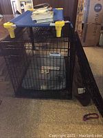 Large black metal wire dog kennel shown with blue elevated dog bed on top, in a carpeted indoor setting.
