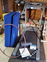 Overall shot: treadmill, three blue mats, hula hoop leaning, black foam roller and grey handheld roller on deck, ankle weights box on floor