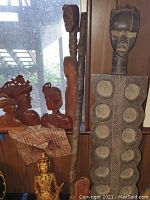 Photo showing various carved wooden artifacts including African wooden bust bookends, tall carved walking stick, and a carved wall hanging panel with circular patterns and a human head top.