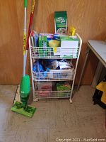 Full view of white 3-tier rolling cart loaded with cleaning supplies including Swiffer broom in front and mop cleaning boxes on top shelf.