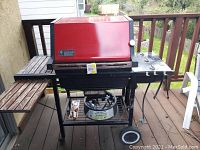View of red Weber gas grill on wooden deck, showing left side wood slatted shelf, propane holder below, and three burner dials on right control panel