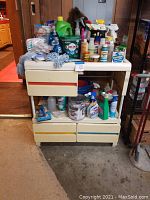 Wide shot of cleaning supplies arranged on top of and inside a plastic utility cart with drawers in a garage setting.