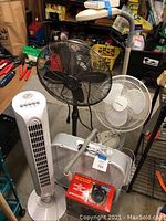 Image of five fans and two white adjustable desk lamps on a basement shelf surrounded by other items.
