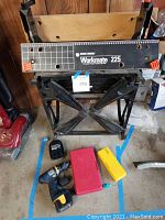 Photo showing Black & Decker Workmate 225 portable project bench folded open with wooden top and orange clamps. Ryobi HP 496 cordless drill with blue and yellow coloring lying on the ground with charger and two plastic drill bit cases in red and yellow.