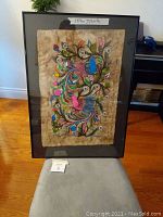 Framed colorful Mexican bird print on textured beige background, standing on a grey chair, showing full front view