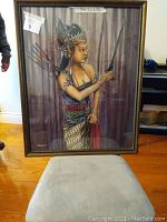 Full frontal image of the framed painting showing an Indonesian woman in traditional attire, holding a dagger with arrows on her back, placed against a curtain background with a beige upholstered surface in front.