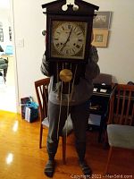 Front view of vintage wall-mounted mechanical chime clock with Roman numeral face, visible pendulum and weights, held by person indoors