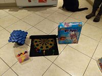 Photo showing Kerplunk game parts including blue plastic stemmed base, marbles container, fabric magnetic dart board with four magnetic darts, and Kerplunk box