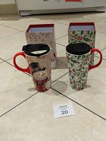 Photo showing two ceramic Christmas mugs with lids and their matching decorative boxes in the background, placed on a tile floor.