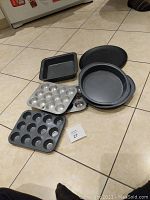 Photo showing a group of baking tins on a tile floor: three mini muffin tins, two round tins, one square tin, and one pizza tin.
