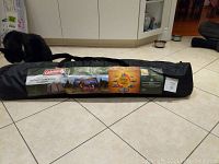 Full view of the Coleman Instant Screened Canopy in carrying bag with label and storage strap visible, laid on tiled floor with a black cat nearby.