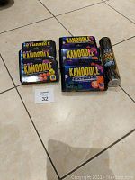 Photo shows 3 Kanoodle classic puzzle game sets stacked on left, 3 Kanoodle Extreme puzzle game sets stacked on right, and PickUp Snakes game in a clear cylindrical container on far right. All items are arranged on a tiled floor.