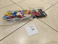 Two plastic bags on a tiled floor containing assorted LEGO bricks in different colors and sizes, with a lot number card visible.