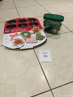Wilton metal cookie shapes pan in packaging and green container of cookie cutters on tile floor.