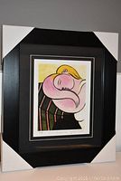 Framed fine art print in black wood frame with matted border showing abstract woman with yellow hair reclining on chair.