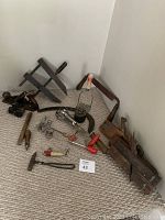 Wide-angle photo showing assortment of antique tools including wooden clamps, hand planes, a vintage fishing lure, hand-crank egg beater, cookie cutter, and small hammer all placed on carpet in corner.