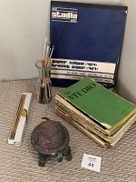 Photo showing Studio art books stacked next to metal wine carafe with artist brushes, Talens tracing paper pad standing upright behind, and Raku pottery clay turtle figurine in front.
