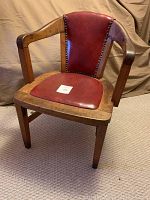 Front left angle view of the antique wooden office chair showing faux-leather padded seat and backrest with decorative nailhead trim and wooden armrests.